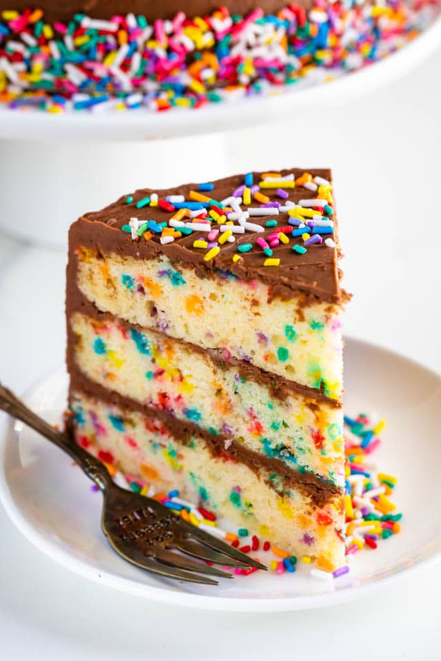 One slice of layered funfetti birthday cake with chocolate frosting and rainbow sprinkles on a white plate with fork