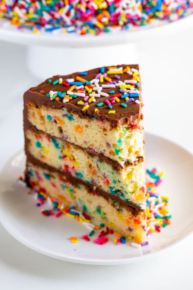 One slice of layered funfetti birthday cake with chocolate frosting and rainbow sprinkles on a white plate with one bite missing