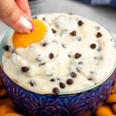 cannoli dip in a blue bowl with orange cookie wafers next to the bowl and hand dipping cookie into the bowl.