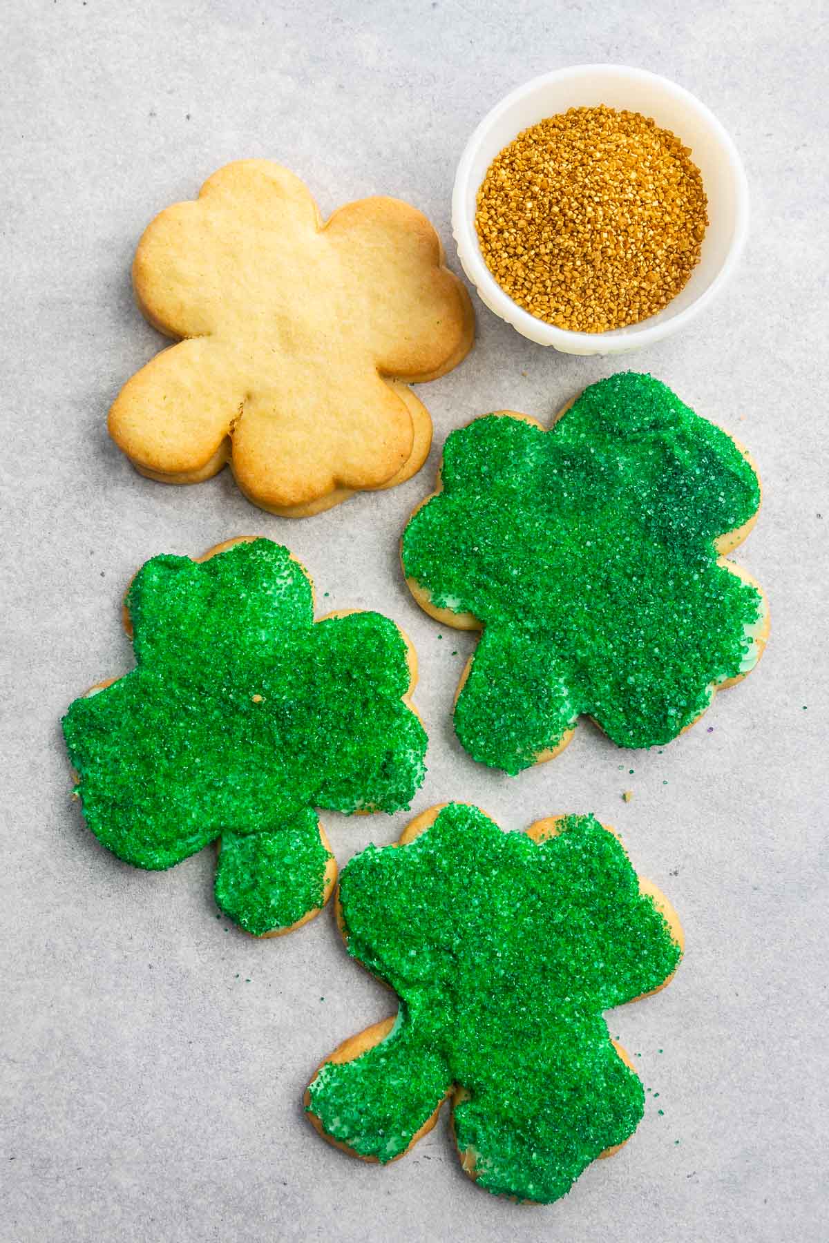 Overhead shot of three decorated shamrock sugar cookies with one undecorated next to gold sprinkles