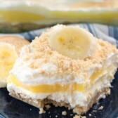 Slice of No Bake Banana Pudding Dream Dessert on a dark blue plate with the rest of the cake in a glass dish in the background